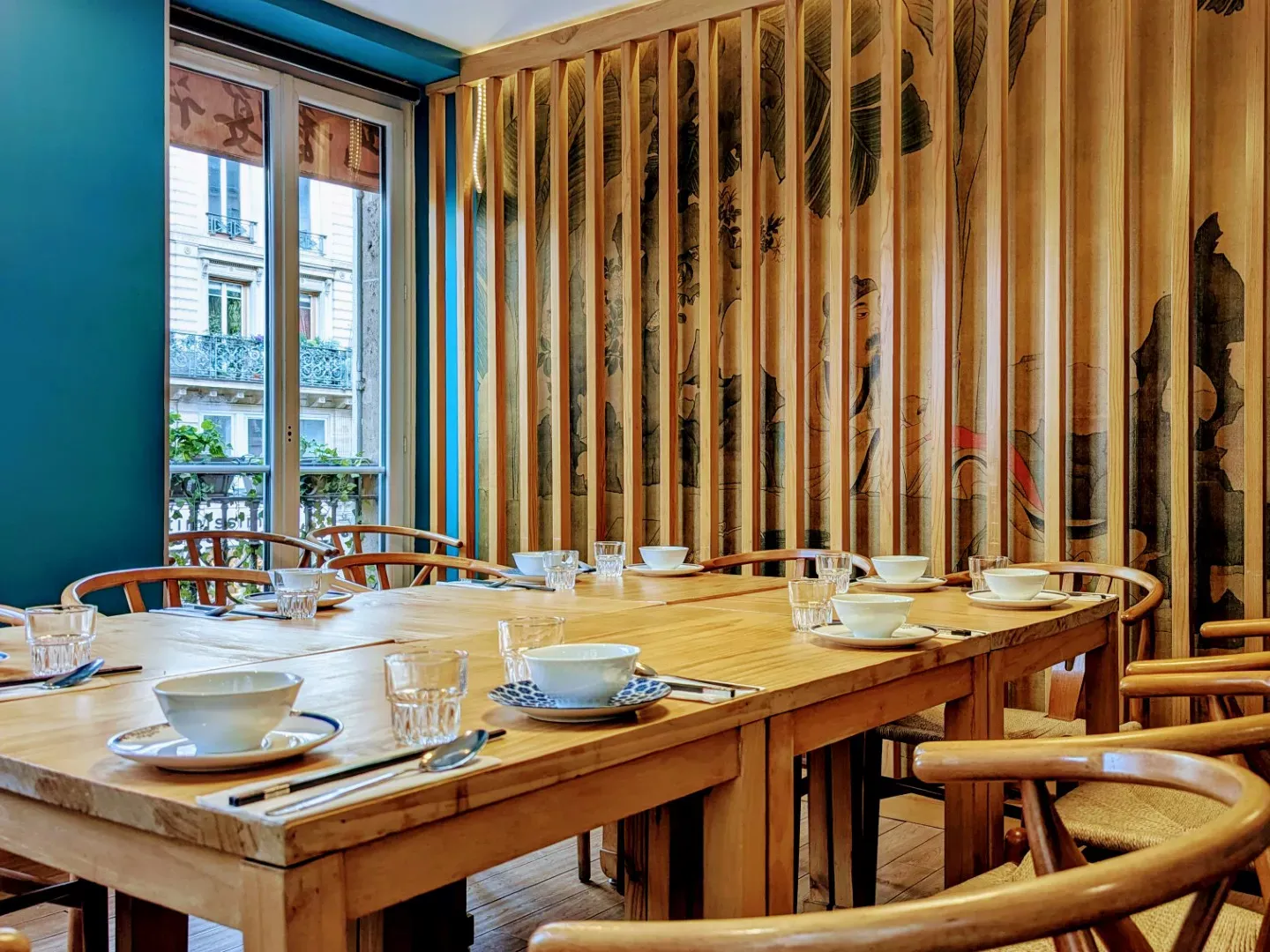 Petite salle avec une table ronde dressée, une porte bleu-gris et une fresque de scènes de rue derrière un treillis de bois